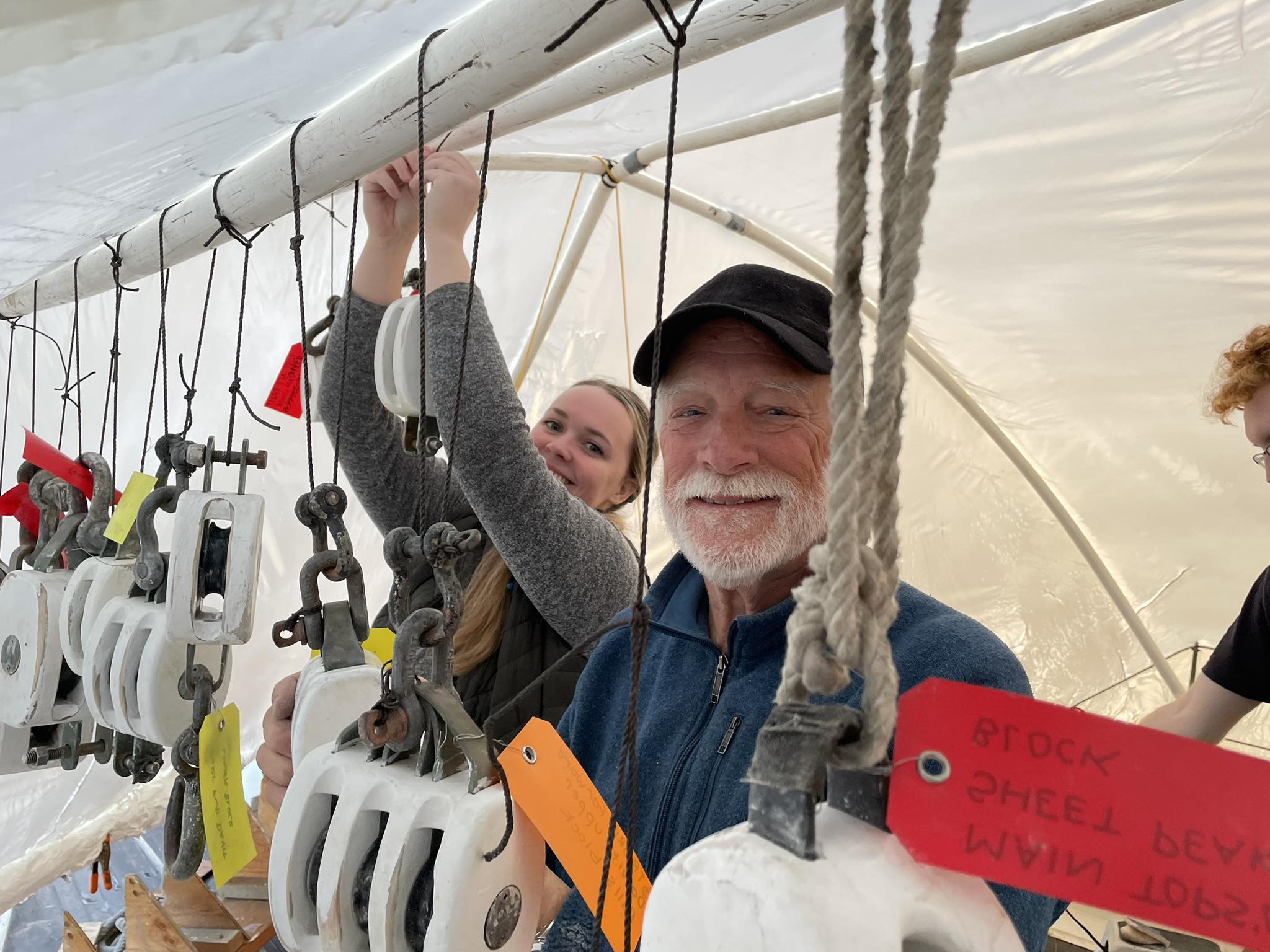 A young woman and an older man in a white beard and wool hat smile as they work under cover aboard Adventuress, volunteering for a work weekend in January restoring blocks.
